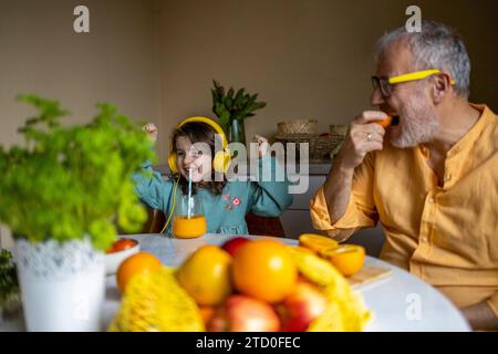 Homme senior mangeant orange et heureuse fille mignonne avec des écouteurs écoutant de la musique et dansant tout en buvant du jus avec de la paille à table Banque D'Images