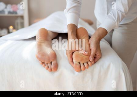 Coupez les mains d'une thérapeute féminine anonyme massant le pied du client allongé sur le lit pour soulager la douleur en appuyant doucement sur les points de pression au salon de spa Banque D'Images
