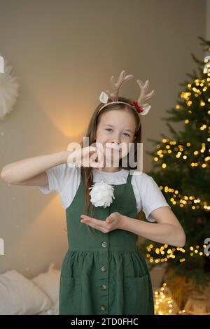 'Une jeune fille joyeuse portant un bandeau de renne fait des gestes d'amour devant un sapin de Noël avec des lumières brillantes.' Banque D'Images