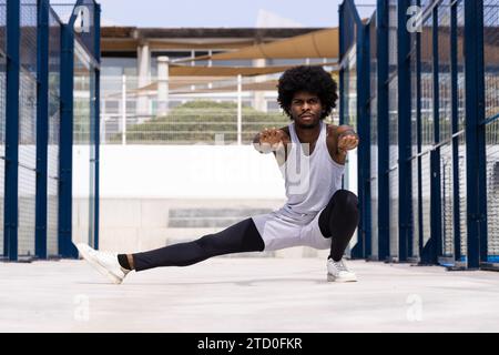 Corps entier de sérieux athlète masculin afro-américain en activewear avec les mains serrées étirant la jambe et faisant accroupi contre la construction en ville Banque D'Images
