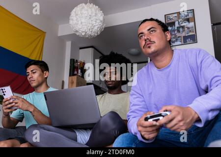 Divers jeune homme messagerie sur smartphone et amis avec ordinateur portable et console jouant au jeu vidéo en ligne et regardant l'écran dans le salon pendant Banque D'Images