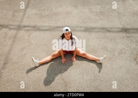 Une jeune femme portant une casquette blanche et une tenue de sport prend une pause et s'assoit sur le sol en béton avec ses jambes écartées Banque D'Images