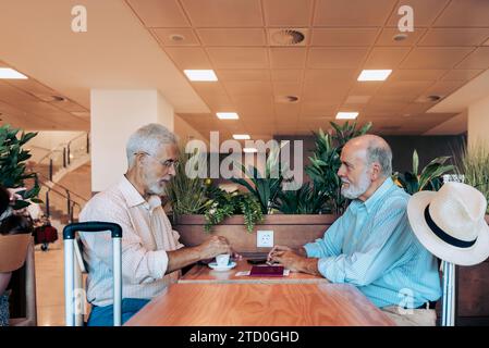 Vue latérale d'amis barbus de sexe masculin habillés dans des occasionnels parlant et prenant un café sur une table en bois dans la cafétéria de l'aéroport Banque D'Images