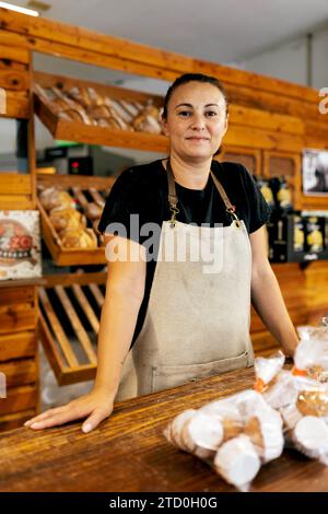 Portrait de vendeuse positive confiante en tablier avec ses mains sur le comptoir debout par des pains fraîchement cuits disposés sur des étagères en bois en bak Banque D'Images