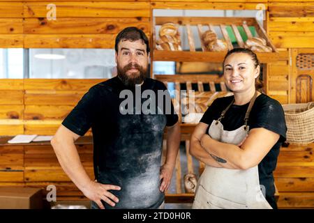 Collègues masculins et féminins positifs dans le tablier et occasionnels regardant la caméra tout en se tenant debout contre des étagères en bois avec des produits cuits dans la boulangerie Banque D'Images