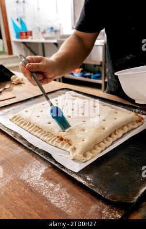 Couper les mains de l'anonyme faire chef tenant la brosse et le bol enduire la pâte farcie avec le jaune d'oeuf dans le plateau tout en faisant la pâtisserie dans la cuisine à la boulangerie Banque D'Images