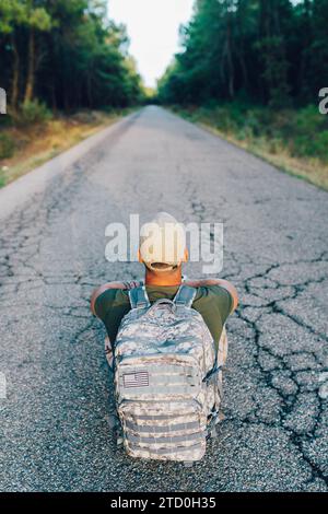 Angle élevé de soldat militaire masculin anonyme portant une casquette avec sac à dos de camouflage assis sur la route contre les arbres dans la forêt Banque D'Images