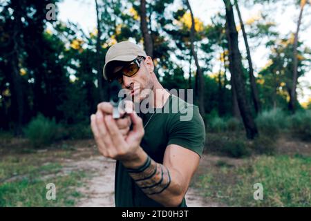 Commando de l'armée de la force spéciale d'âge moyen en casquette et lunettes regardant la caméra tout en visant avec un fusil dans la forêt contre le coucher du soleil Banque D'Images