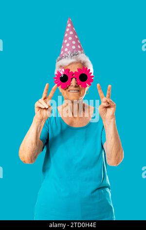 Femme âgée souriante portant des lunettes de soleil roses drôles et une casquette d'anniversaire tout en montrant le geste de paix sur fond bleu Banque D'Images