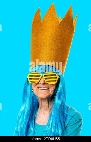 Joyeuse dame senior portant perruque bleue, grande couronne et lunettes de soleil rayées jaunes souriant à la caméra sur fond bleu Banque D'Images