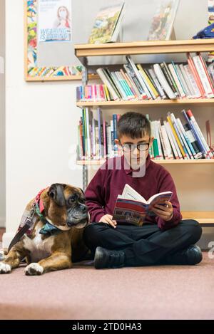 Un jeune étudiant masculin est assis sur le sol de la bibliothèque de l'école lisant un livre au chien d'apprentissage / de thérapie de l'école. Banque D'Images