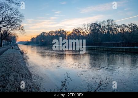 Une scène tranquille tôt le matin où un lever de soleil doux orne le ciel de couleurs pastel au-dessus d'un canal glacé. Les arbres, dépourvus de feuilles, se présentent comme des témoins silencieux du début de la journée, leurs branches créant des motifs délicats contre le ciel illuminé. Le canal, partiellement gelé, reflète les pastels calmes du ciel et les contours sombres des arbres. Une traînée de vapeur d'un avion ajoute un élément moderne au paysage par ailleurs intemporel, faisant allusion à l'éveil du monde. Lever de soleil serein sur le canal glacé. Photo de haute qualité Banque D'Images