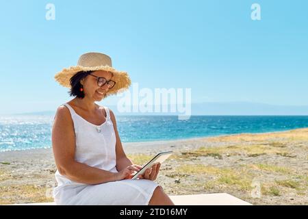 Femme mature portant un chapeau de paille en utilisant une tablette numérique à la plage. Femme d'âge moyen utilisant un appareil numérique pour faire des achats en ligne, des banques, des médias sociaux o Banque D'Images