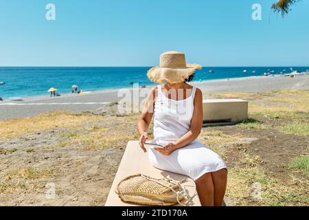 Femme mature portant un chapeau de paille en utilisant une tablette numérique à la plage. Femme d'âge moyen utilisant un appareil numérique pour faire des achats en ligne, des banques, des médias sociaux o Banque D'Images