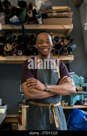 Un jeune homme se tient debout dans son atelier de cuir où il conçoit et fabrique des articles de mode tels que des sacs suspendus, des portefeuilles et des ceintures. Banque D'Images