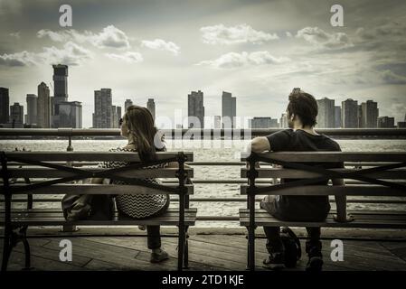 Un jeune homme et une femme se détendant sur des bancs à Battery Park, avec Jersey City Skyline en arrière-plan - Manhattan, New York City Banque D'Images