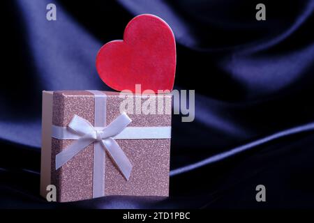 Boîte cadeau avec noeud noué et coeur rouge sur fond tissu de soie noir Banque D'Images