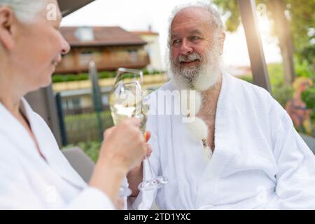 Couple senior dans des peignoirs blancs serrant des verres de champagne, homme regardant la femme à l'hôtel spa Resort Banque D'Images
