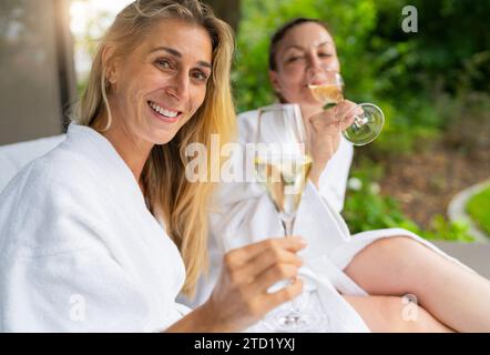 Deux femmes souriantes vêtues de peignoirs blancs dégustant du champagne à l'extérieur sur une chaise longue dans un centre thermal Banque D'Images