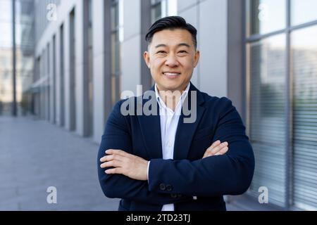 Portrait rapproché d'un jeune homme d'affaires souriant debout dans un costume à l'extérieur d'un centre de bureau, croisant ses bras sur sa poitrine et regardant avec confiance Banque D'Images