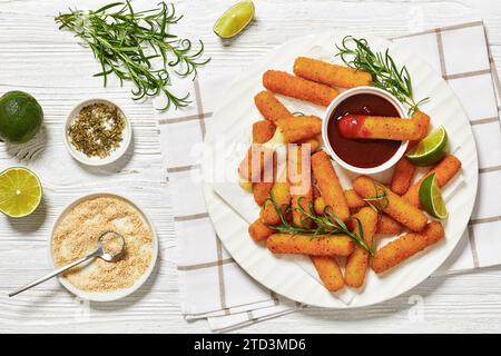 bâtonnets de mozzarella panés frits servis avec sauce, tranches de citron vert et romarin frais sur un plateau blanc sur une table en bois blanc avec des ingrédients, hor Banque D'Images