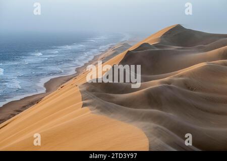 Dunes de sable et mer dans la lumière du soir, le vent souffle du sable sur les dunes, vue sur la côte atlantique depuis les hautes dunes de sable, Sandwich Harbour, Namib Banque D'Images