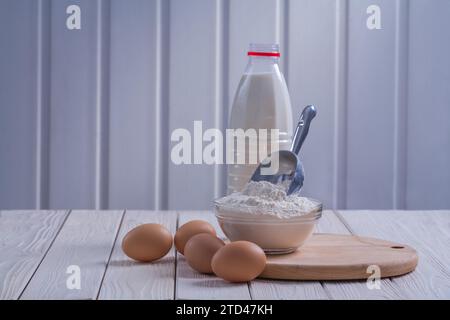 Vue horizontale oeufs farine dans le bol cuillère bouteille de lait sur blanc laqué vieux panneau en bois concept de nourriture et de boisson Banque D'Images