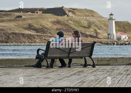 Couple assis sur un banc sur la promenade riveraine de Halifax, avec le phare de George Island en arrière-plan Banque D'Images