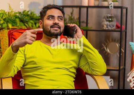 Appel téléphonique, bonnes nouvelles, commérages. Heureux surpris homme indien ayant une conversation agréable sur smartphone, appréciant parler avec un ami à la chambre moderne de la maison. Gars arabe appréciant téléphone portable parlant Banque D'Images