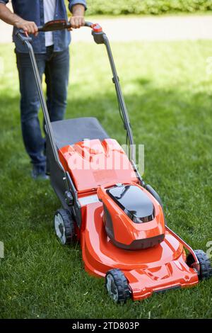 Travailleur anonyme masculin tondant la pelouse de jardin avec un nouveau coupe-herbe sans fil. Vue du dessus de la tondeuse à gazon orange brillant moderne, actionnée par un bricoleur, on Banque D'Images