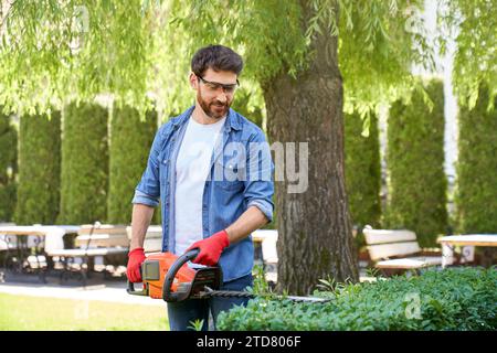 Homme caucasien dans des lunettes de protection coupant buisson de buis avec taille-haie à l'extérieur. Vue de face du jardinier professionnel en utilisant des équipements modernes, tandis que Banque D'Images