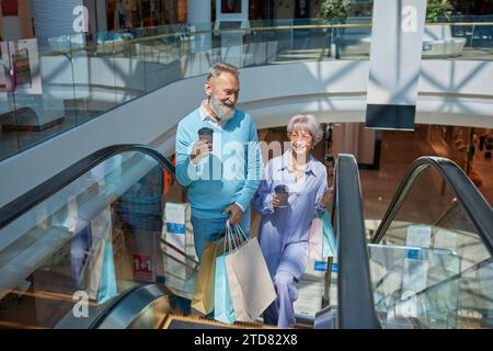 Couple de personnes âgées descendant l'escalator du centre commercial en appréciant ensemble Banque D'Images