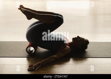 Fille mince faisant du yoga avec un gros rouleau de massage sur le sol. Banque D'Images