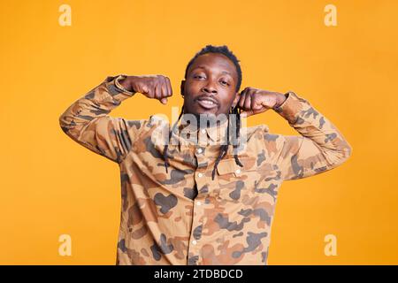 Homme confiant athlétique fléchissant les muscles des bras entraînés devant la caméra en studio sur fond jaune. Jeune afro-américain appréciant des exercices sportifs, s'exerçant à la force du biceps. Banque D'Images