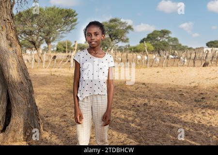 Village, fille africaine à la ferme, kraal avec des chèvres en arrière-plan, Kalahari petit bétail Banque D'Images