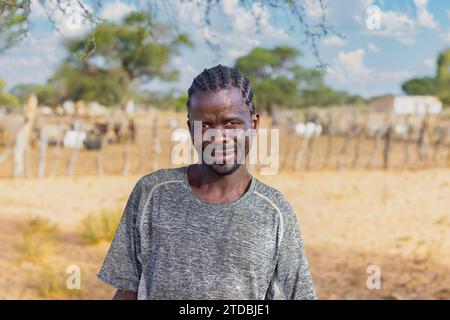 Village, homme africain avec des dreadlocks à la ferme, kraal avec des chèvres en arrière-plan, Kalahari petit bétail Banque D'Images