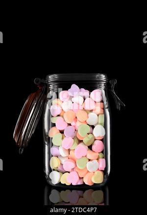 Un pot de rangement ou de mise en conserve en verre rempli de coeurs Valentines Candy isolés sur noir avec reflet, avec couvercle ouvert. Banque D'Images