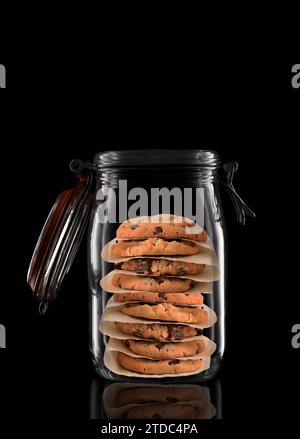 Un pot de stockage ou de mise en conserve en verre avec des cookies aux pépites de chocolat isolés sur noir avec réflexion, avec le couvercle ouvert. Banque D'Images