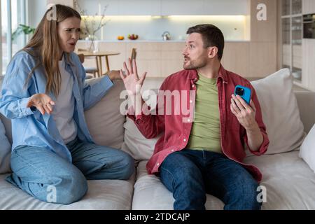 Jaloux en colère mari méfiant exige mot de passe de la femme téléphone portable assis sur le canapé à la maison. Banque D'Images