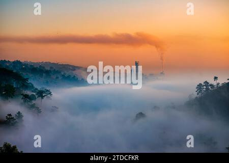 La brume tôt le matin couvre le paysage, tandis que la fumée épaisse s'écoule et dérive d'une cheminée dans une usine à Umiam, Meghalaya, en Inde. Banque D'Images