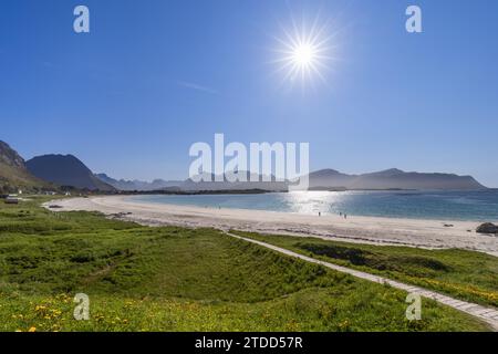 Sentier ensoleillé serpentant à travers l'herbe et les fleurs sauvages jusqu'au sable blanc de Rambergstranda Beach dans la baie de Jusnesvika, Lofoten, avec une mer scintillante A. Banque D'Images