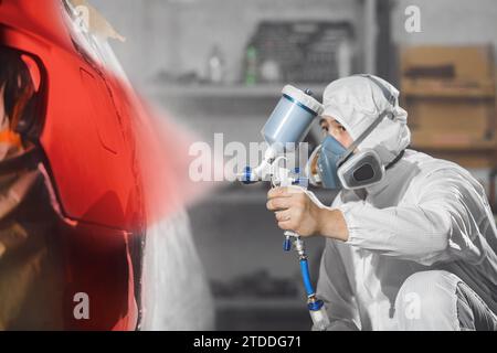 Pulvériser le peintre ouvrier dans un gant de protection avec la peinture rouge pulvérisateur d'aérographe à la carrosserie de la voiture dans la chambre de peinture. Banque D'Images