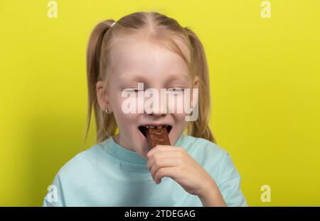 Une belle fille joyeuse aux cheveux blancs de sept ans mange une barre de chocolat sur fond jaune. Les enfants sont des amoureux de la malbouffe sucrée, horizonto Banque D'Images