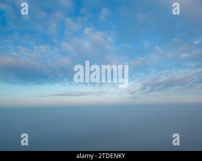Cette photographie, prise avec un drone au-dessus de la brume, présente une vue à couper le souffle semblable à être au-dessus du niveau des nuages. La brume couvre le paysage, créant un horizon sans couture où le ciel se confond avec la terre dans une étendue bleue tranquille. Le ciel est orné de délicats nuages d'altocumulus, teintés des doux pastels de l'aube, suggérant le début en douceur d'une nouvelle journée. L'image donne un sentiment de paix et d'isolement, haut au-dessus du monde obscurci et éveillé ci-dessous. Au-dessus des nuages : vue aérienne d'un paysage recouvert de brume à l'aube. Photo de haute qualité Banque D'Images