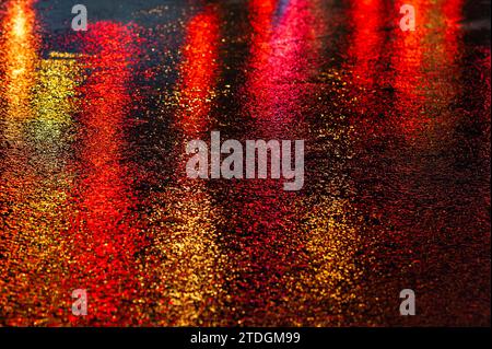 Route asphaltée humide abstraite illuminée de couleur la nuit par des feux arrière de voiture rouges et des feux de signalisation. Banque D'Images