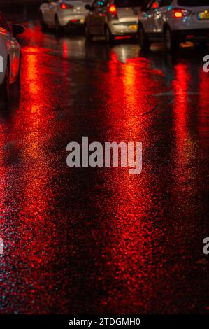 Route asphaltée humide abstraite illuminée de couleur la nuit par des feux arrière de voiture rouges et des feux de signalisation. Banque D'Images