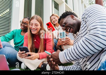Groupe de jeunes lycéens multiethniques souriants utilisant des téléphones cellulaires. Les gars et les femmes regardent les médias sociaux avec leurs smartphones, tout en profitant ensemble assis sur la pelouse du campus universitaire. Photo de haute qualité Banque D'Images