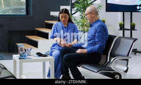 Assistante asiatique prenant un échantillon de sang mesurant le taux de sucre pour faire un test d'insuline avec glucomètre pour patient âgé diabétique pendant la consultation. Le contrôle infirmier de la glycémie en salle d'attente hospitalière Banque D'Images