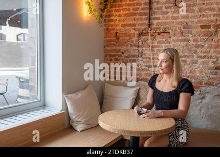 Cette image capture un moment serein alors qu'une jeune femme est assise près d'une fenêtre dans un café confortable, tenant un smartphone dans ses mains. L'intérieur allie design contemporain et charme rustique, avec des murs en briques apparentes et un éclairage chaleureux qui complètent sa tenue moderne. La fenêtre permet à la lumière naturelle de filtrer, ajoutant une lueur douce au cadre, ce qui est à la fois accueillant et tranquille. Son expression est réfléchie, suggérant une pause tranquille dans une journée bien remplie. Ambiance contemplative dans un café rustique. Photo de haute qualité Banque D'Images