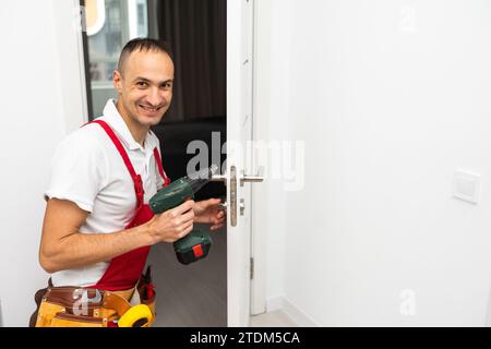 Serrurier joyeux l'installation d'une serrure de porte sur une nouvelle porte blanche et regardant la caméra Banque D'Images